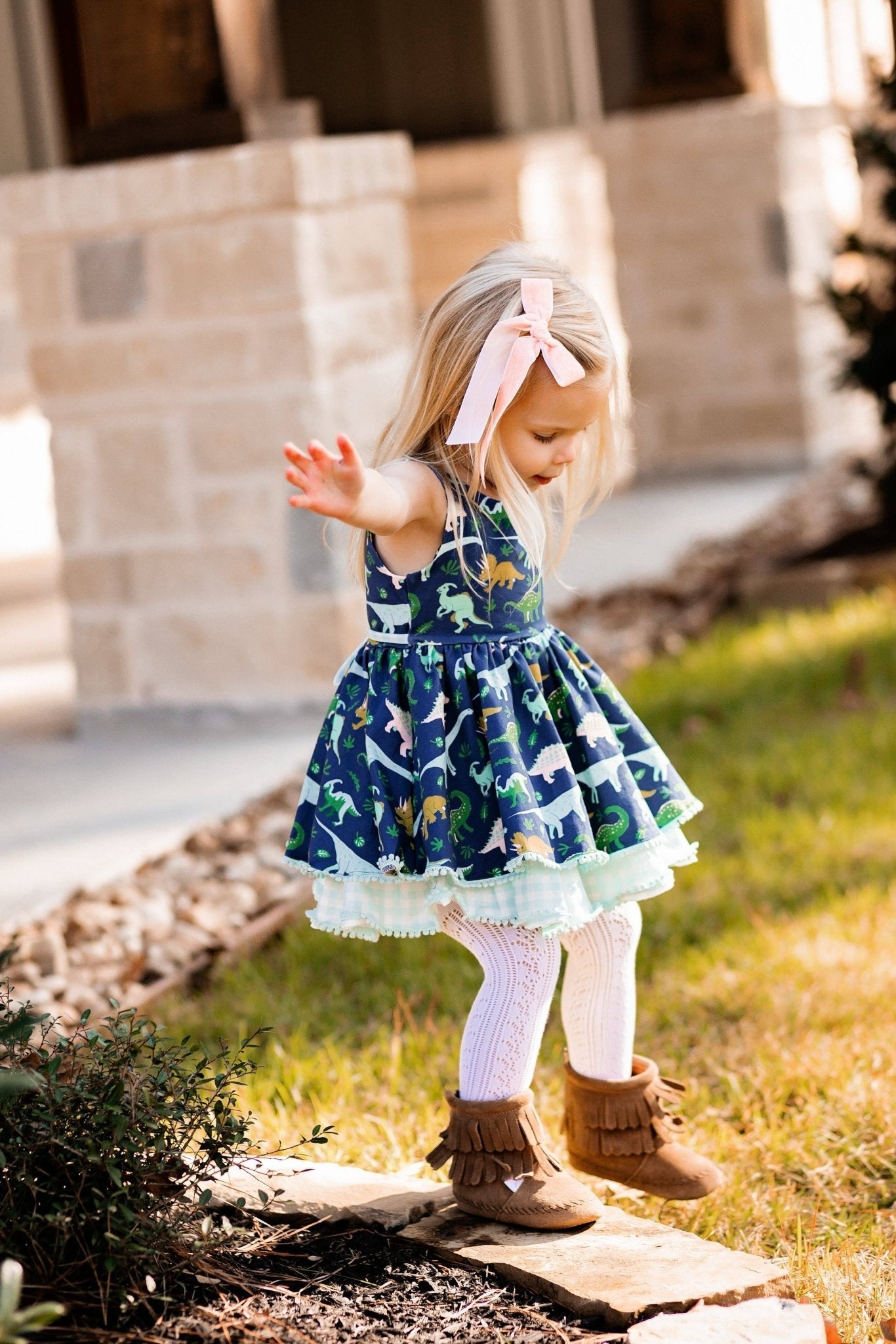 Young girl in a patterned dress with a pink bow standing outdoors.
