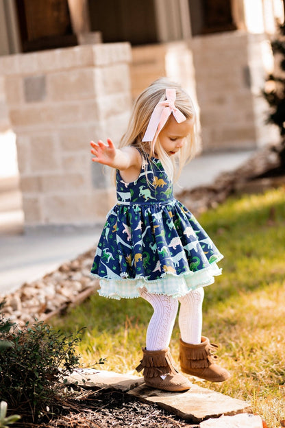 Young girl in a patterned dress with a pink bow standing outdoors.