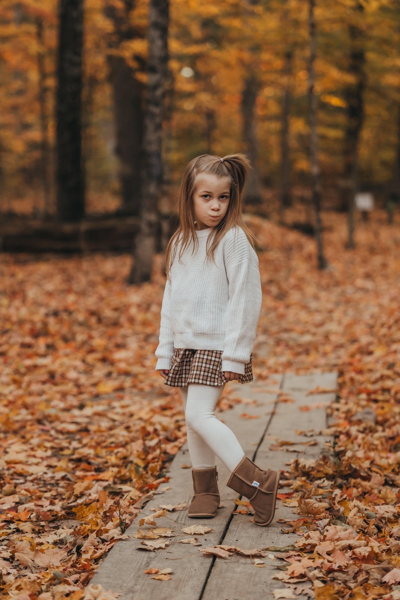 Young girl in a white sweater and plaid skirt walking on a wooden path in a forest with autumn leaves.